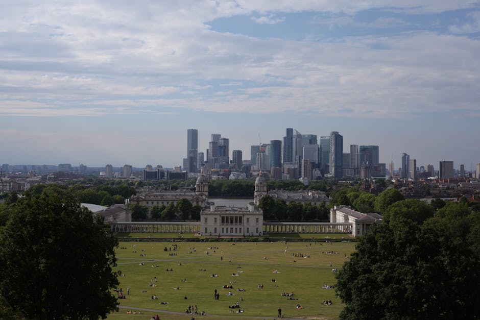 A wide view of a historic palace with white classical architecture, featuring symmetrical wings, central pediment, and domed towers on either side, situated behind a large open grassy park with pathwa