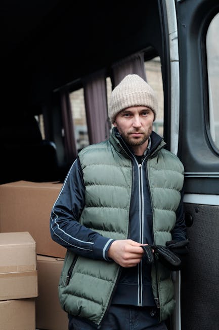 A man sitting on the pavement next to an open moving van loaded with stacked cardboard boxes of various sizes, some sealed with packing tape. The man is wearing a black cap, white t-shirt, and blue je