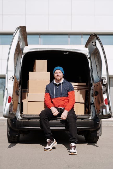A man with a beard, wearing a red cap backwards, a beige and brown jacket over a checked shirt, and grey gloves, stands inside the back of a van loaded with cardboard moving boxes. The van's interior 